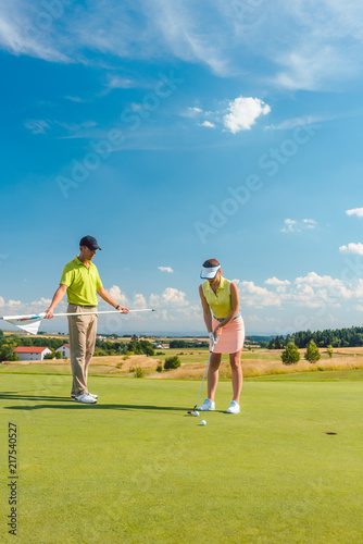 Full length of a woman calculating the trajectory of the ball to the hole, while playing professional golf with her male match partner or instructor outdoors in summer