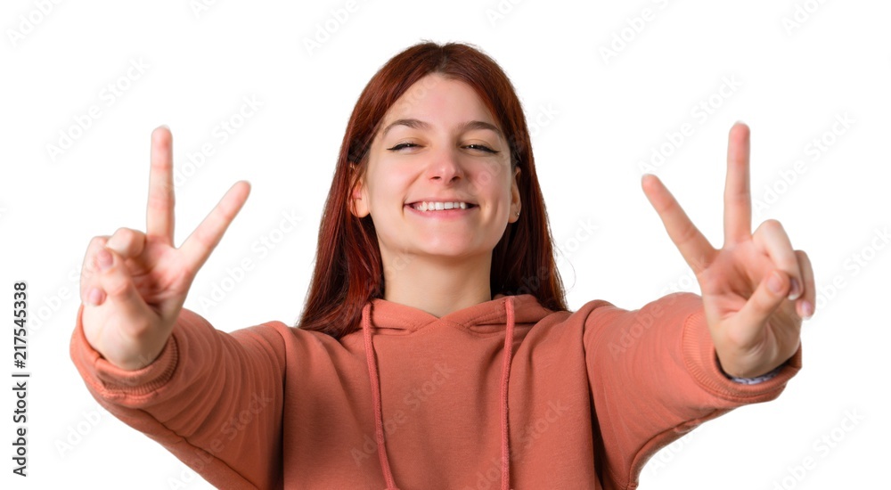 Young redhead girl with pink sweatshirt smiling and showing victory sign with both hands and with a cheerful face on isolated white background Young redhead girl with pink sweatshirt smiling and showing victory sign with both hands and with a cheerful face on isolated white background