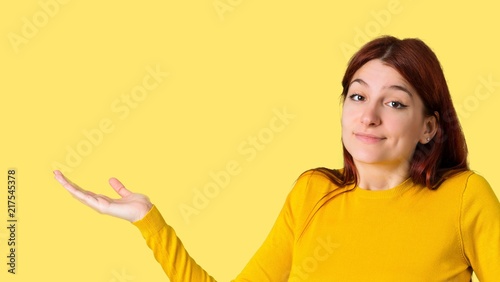 Young redhead girl with yellow sweater making unimportant and doubts gesture while lifting the shoulders and the palms of the hands on isolated yellow background