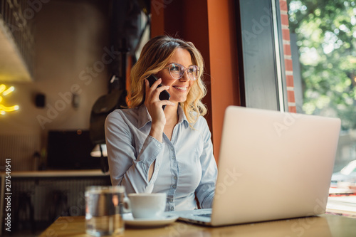 Portrait of young business woman with glasses sitting in cafe in front of her laptop and talking on mobile phone