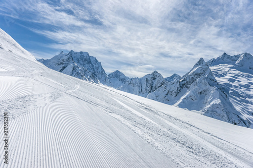 Ski slope with the mountains Dombay-Ulgen, Sofrudzhu and peak Ine on the horizon in winter sunny day. Dombai ski resort, Western Caucasus, Russia.