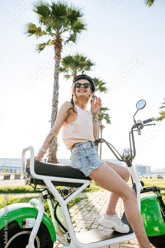 people, transport, technology, travel and vacation concept - outdoor portrait of pretty cute tourist woman smiling beside her electric bike in park with palms in summer morning. photo