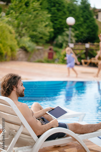 Positive freelance man using laptop near pool working online, distance work, while his family having fun on background photo