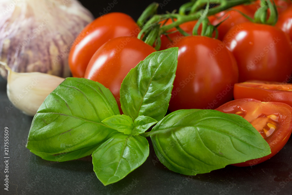 Closeup view of shrubs tomatoes with a leaf of basil, garlic, clove of garlic and sliced tomato lying on the black stone table. Closeup view of shrubs tomatoes with a leaf of basil, garlic, clove of garlic and sliced tomato lying on the black stone table.