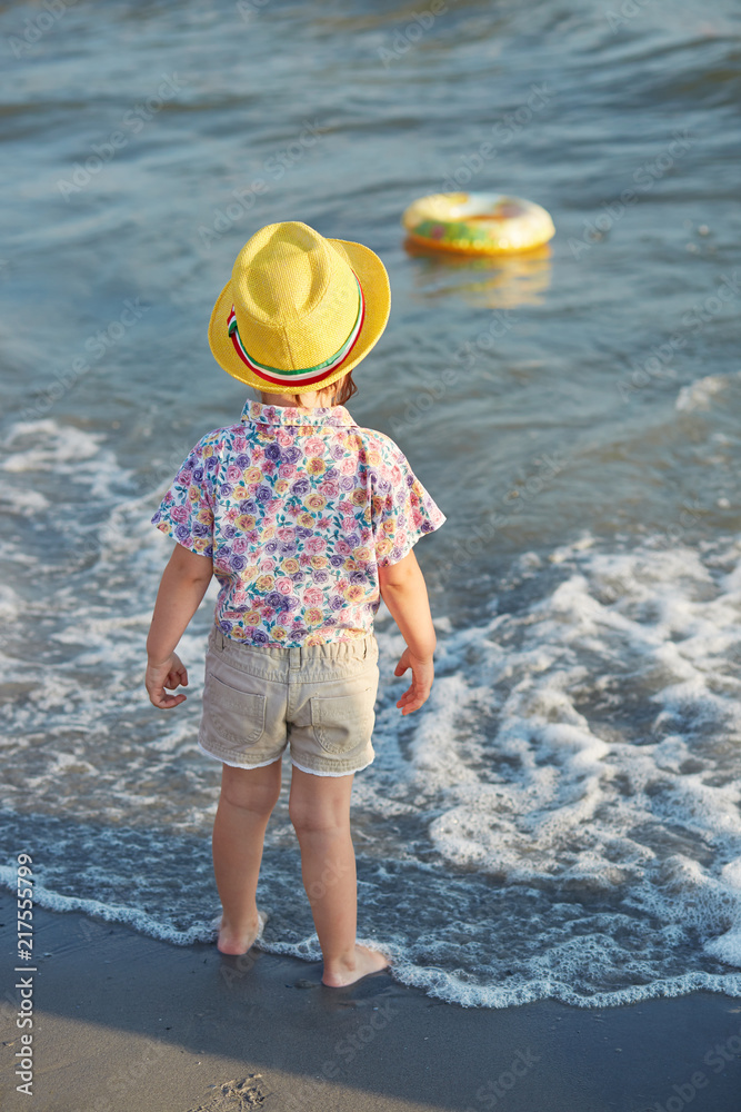 Vertical shot of small fashionable girl stands back, wears summer hat, blouse and shorts, admires sea, plays alone, breathes fresh air. Little child enjoys summer vacation. Childhood concept