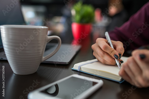 Selective focus on woman's hand with pen records planned schedule in paper notepad sitting at table with cup of coffee and smartphone. Organization of things to do. Blogger writing creative ideas