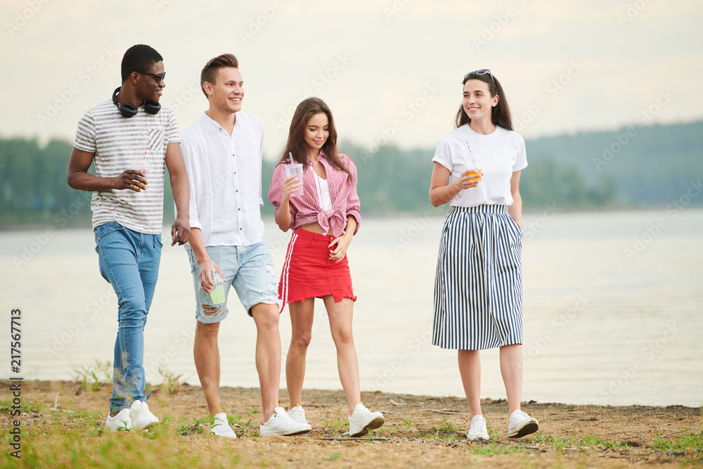 Group of stylish young people with drinks walking along the lake and communicating with each other Group of stylish young people with drinks walking along the lake and communicating with each other
