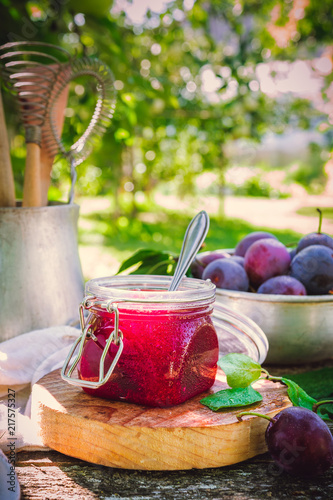 Glass jar with plum jam confiture and ripe plum berries in a basket on a wooden vintage table in the garden with a copy space, the idea of home canning and organic ecological bio nutrition