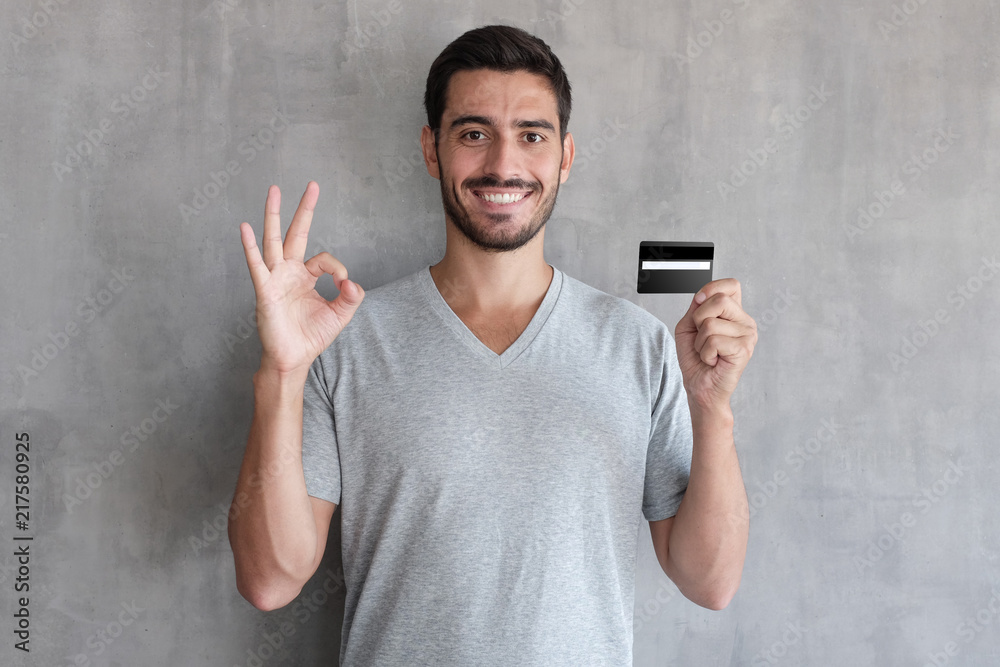 Portrait of young smiling man in t shirt, holding credit card and showing okay sign, standing against gray textured wall Portrait of young smiling man in t shirt, holding credit card and showing okay sign, standing against gray textured wall