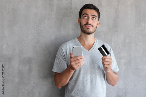 Young handsome man thinking about online shopping , wearing gray t shirt, standing against textured wall with copy space, holding credit card and cell phone