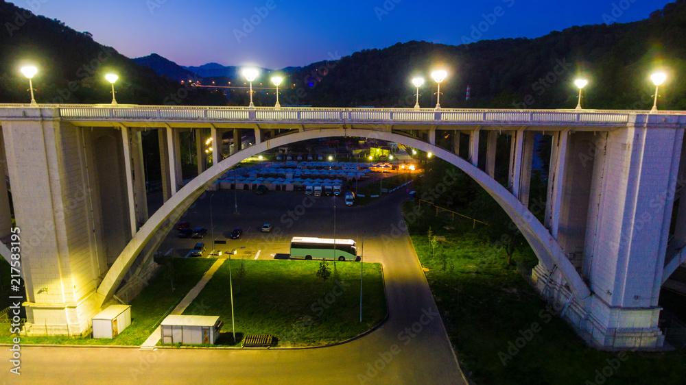 Drone view of the illuminated Matsesta viaduct and the parking under it on the background of mountains at twilight, Sochi, Russia
