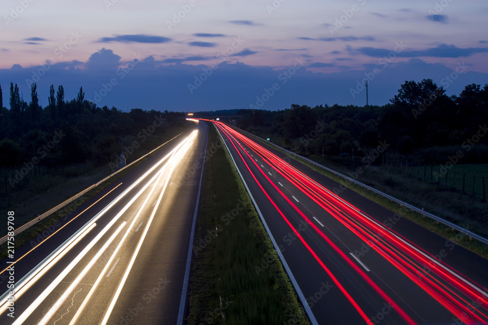 Speedway at night, car lights and road with sky and clouds.