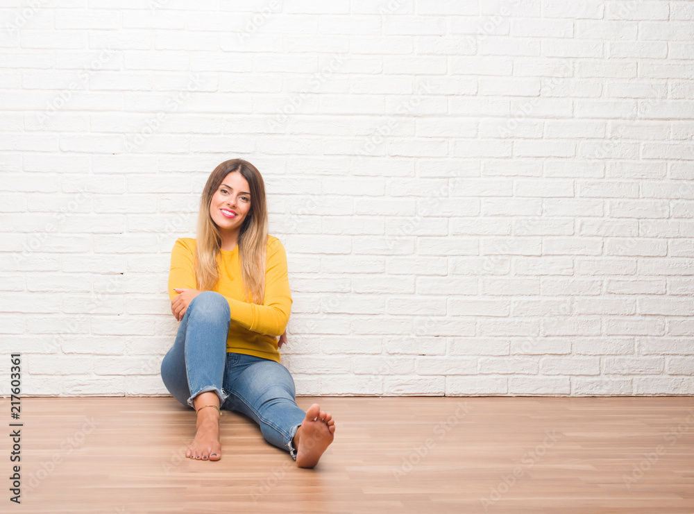 Young adult woman sitting on the floor over white brick wall happy face smiling with crossed arms looking at the camera. Positive person. Young adult woman sitting on the floor over white brick wall happy face smiling with crossed arms looking at the camera. Positive person.