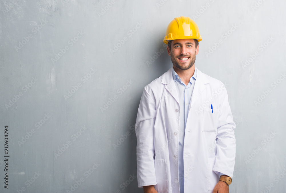 Handsome young engineer man over grey grunge wall wearing safety helmet with a happy face standing and smiling with a confident smile showing teeth Handsome young engineer man over grey grunge wall wearing safety helmet with a happy face standing and smiling with a confident smile showing teeth