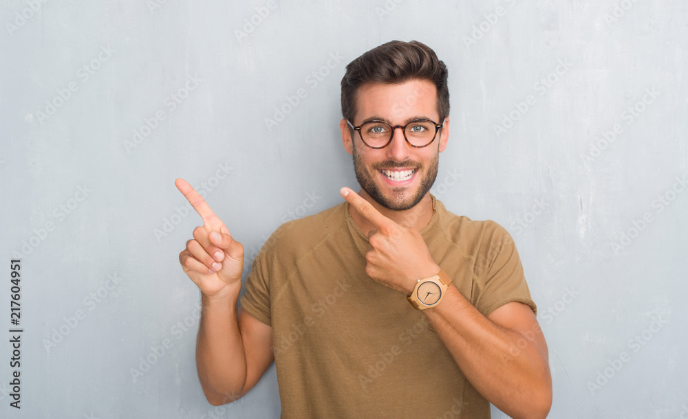 Handsome young man over grey grunge wall wearing glasses smiling and looking at the camera pointing with two hands and fingers to the side. Handsome young man over grey grunge wall wearing glasses smiling and looking at the camera pointing with two hands and fingers to the side.