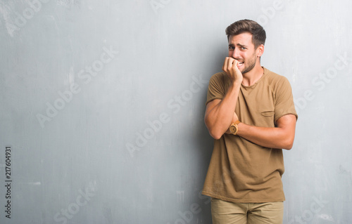 Handsome young man over grey grunge wall looking stressed and nervous with hands on mouth biting nails. Anxiety problem.