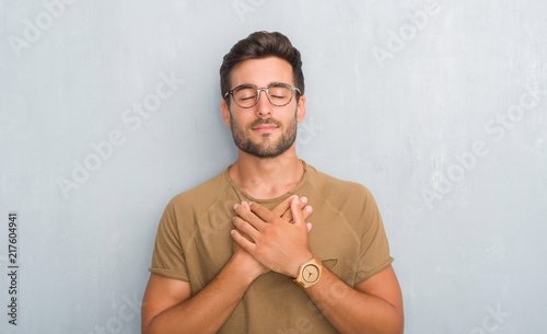 Handsome young man over grey grunge wall wearing glasses smiling with hands on chest with closed eyes and grateful gesture on face. Health concept. photo