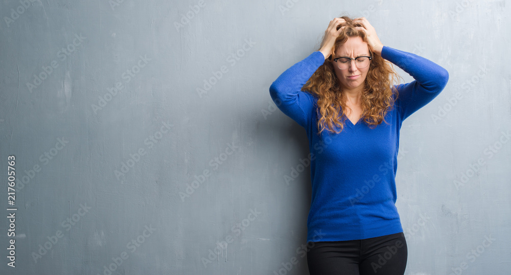 Young redhead woman over grey grunge wall suffering from headache desperate and stressed because pain and migraine. Hands on head.