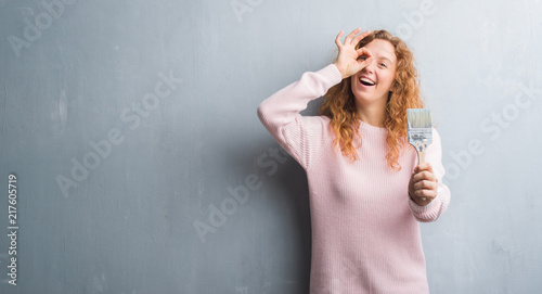 Young redhead woman over grey grunge wall holding paint brush with happy face smiling doing ok sign with hand on eye looking through fingers