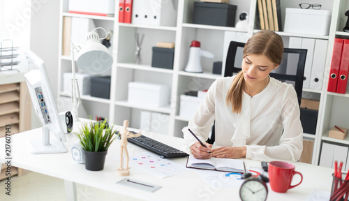 A young girl sits at a table in the office and writes with a pencil in a notebook.