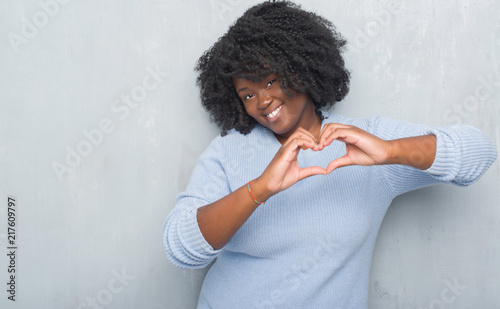 Young african american woman over grey grunge wall wearing winter sweater smiling in love showing heart symbol and shape with hands. Romantic concept. © Krakenimages.com