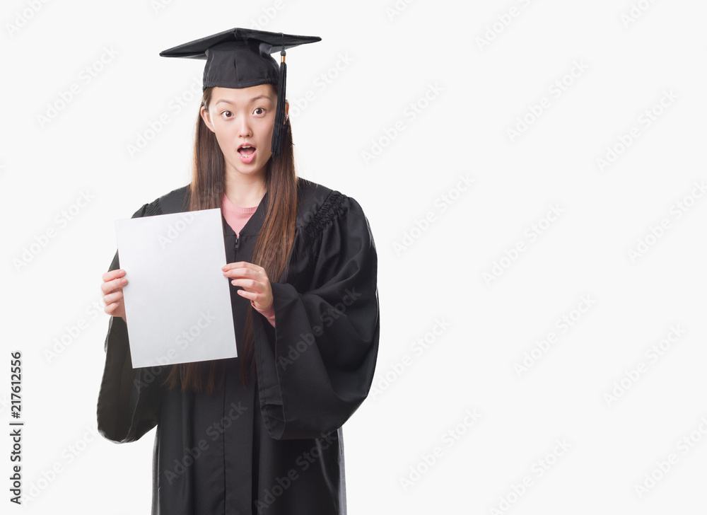 Young Chinese woman wearing graduate uniform holding paper degree scared in shock with a surprise face, afraid and excited with fear expression Young Chinese woman wearing graduate uniform holding paper degree scared in shock with a surprise face, afraid and excited with fear expression