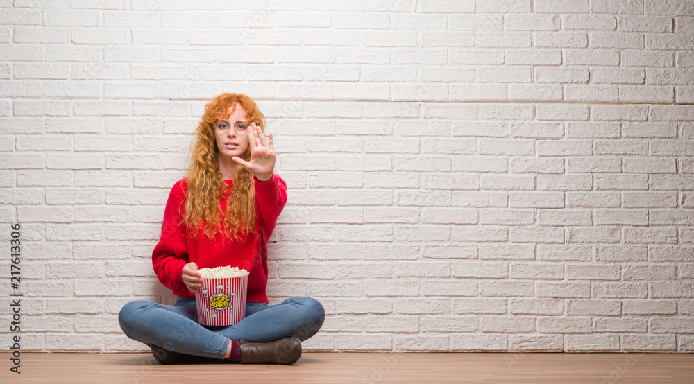 Young redhead woman sitting over brick wall eating popcorn with open hand doing stop sign with serious and confident expression, defense gesture Young redhead woman sitting over brick wall eating popcorn with open hand doing stop sign with serious and confident expression, defense gesture