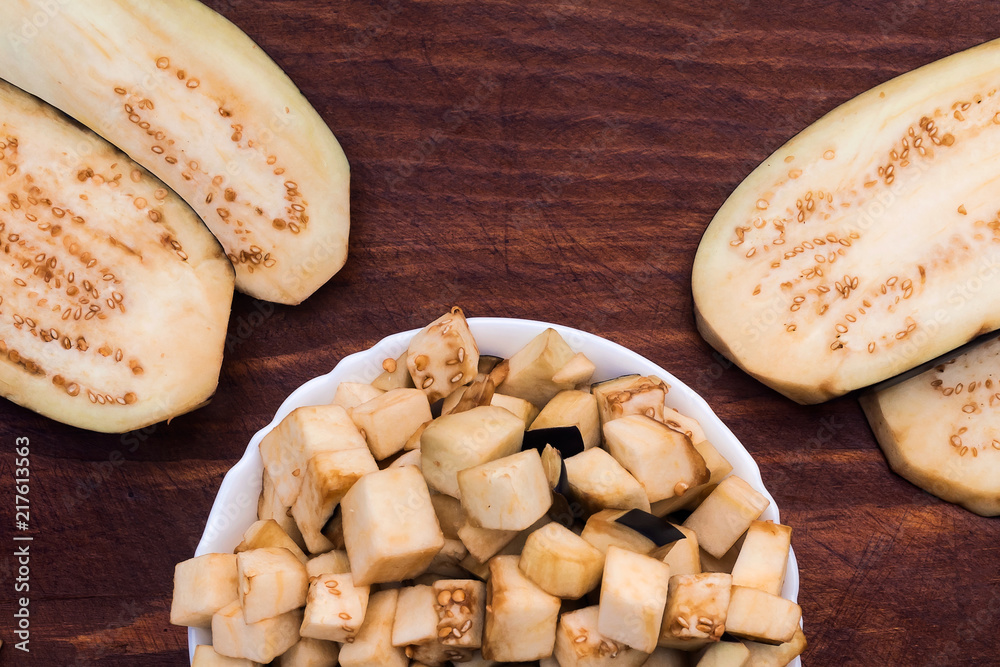 Cubes of sliced eggplant in a white porcelain plate next to wide slices with small seeds, all on a wooden background. Cubes of sliced eggplant in a white porcelain plate next to wide slices with small seeds, all on a wooden background.