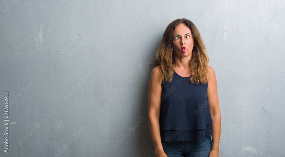 Middle age hispanic woman standing over grey grunge wall making fish face with lips, crazy and comical gesture. Funny expression.