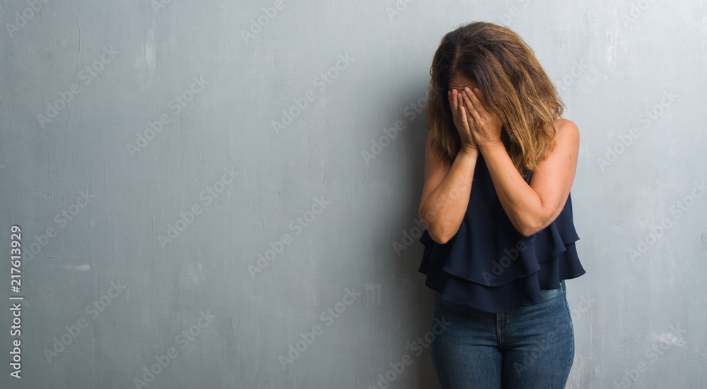 Middle age hispanic woman standing over grey grunge wall with sad expression covering face with hands while crying. Depression concept.