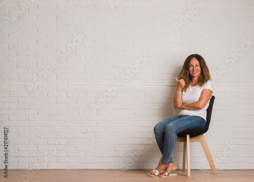 Middle age hispanic woman sitting on chair over white brick walll happy with big smile doing ok sign, thumb up with fingers, excellent sign