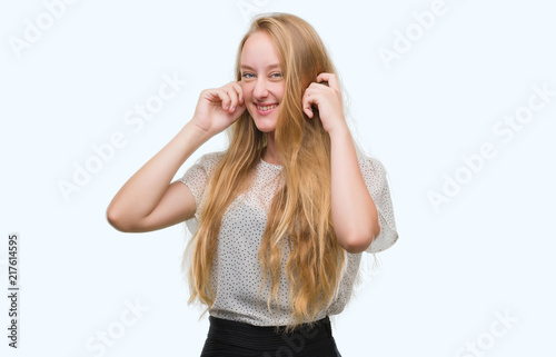 Blonde teenager woman wearing moles shirt covering ears with fingers with annoyed expression for the noise of loud music. Deaf concept.