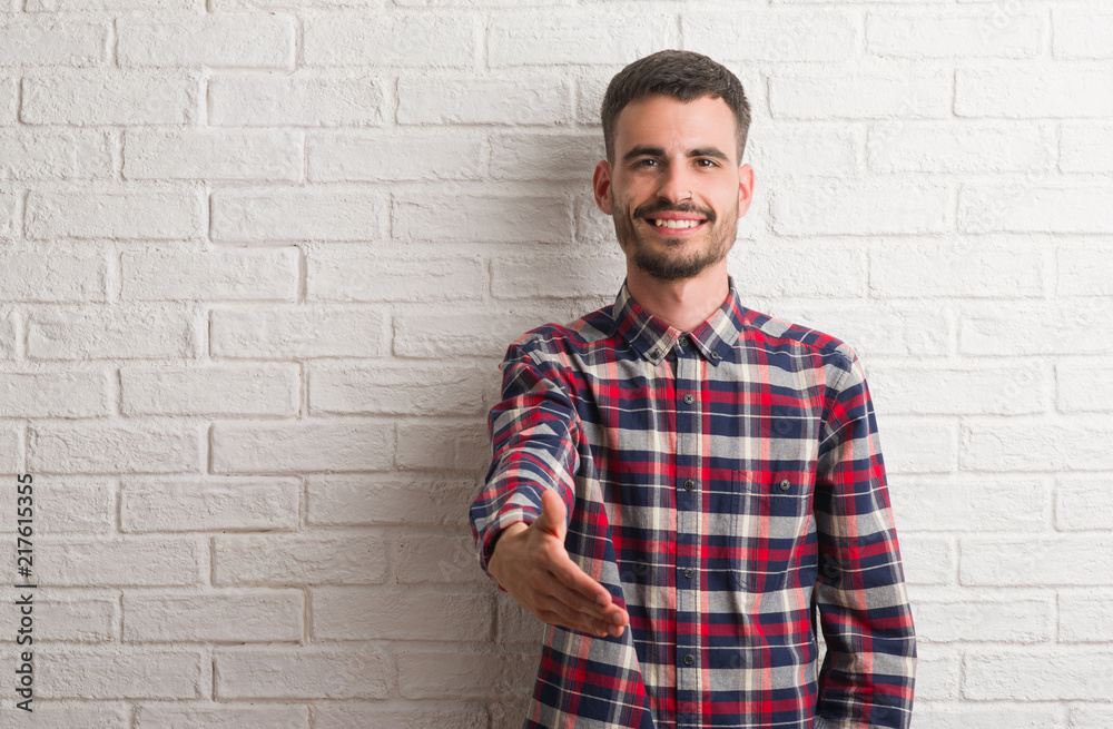 Young adult man standing over white brick wall smiling friendly offering handshake as greeting and welcoming. Successful business. Young adult man standing over white brick wall smiling friendly offering handshake as greeting and welcoming. Successful business.