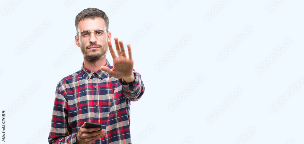 Young hipster adult man holding smartphone with open hand doing stop sign with serious and confident expression, defense gesture Young hipster adult man holding smartphone with open hand doing stop sign with serious and confident expression, defense gesture