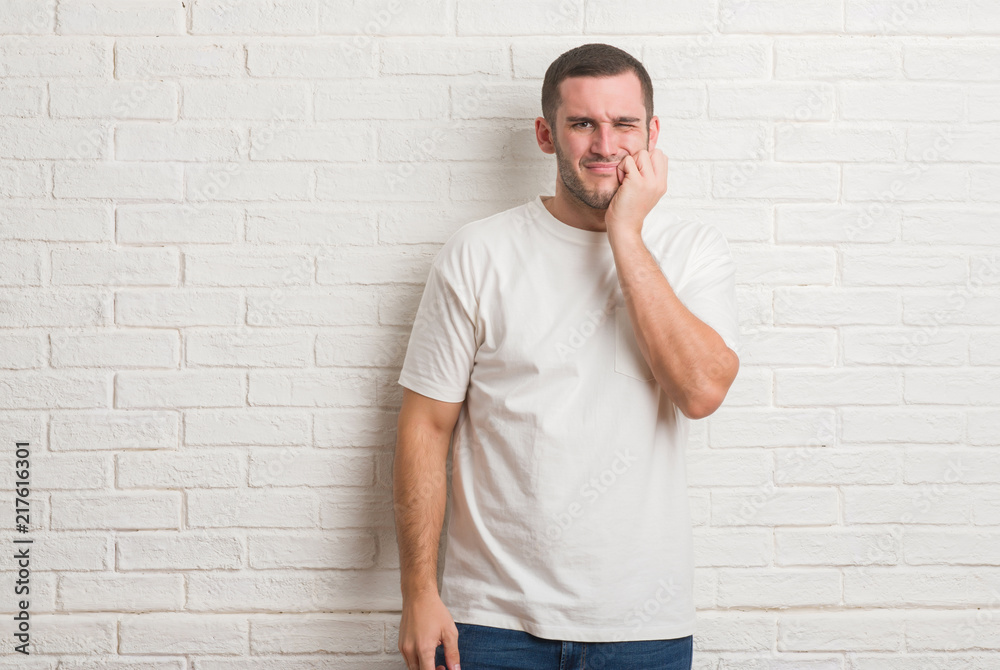 Young caucasian man standing over white brick wall touching mouth with hand with painful expression because of toothache or dental illness on teeth. Dentist concept. Young caucasian man standing over white brick wall touching mouth with hand with painful expression because of toothache or dental illness on teeth. Dentist concept.
