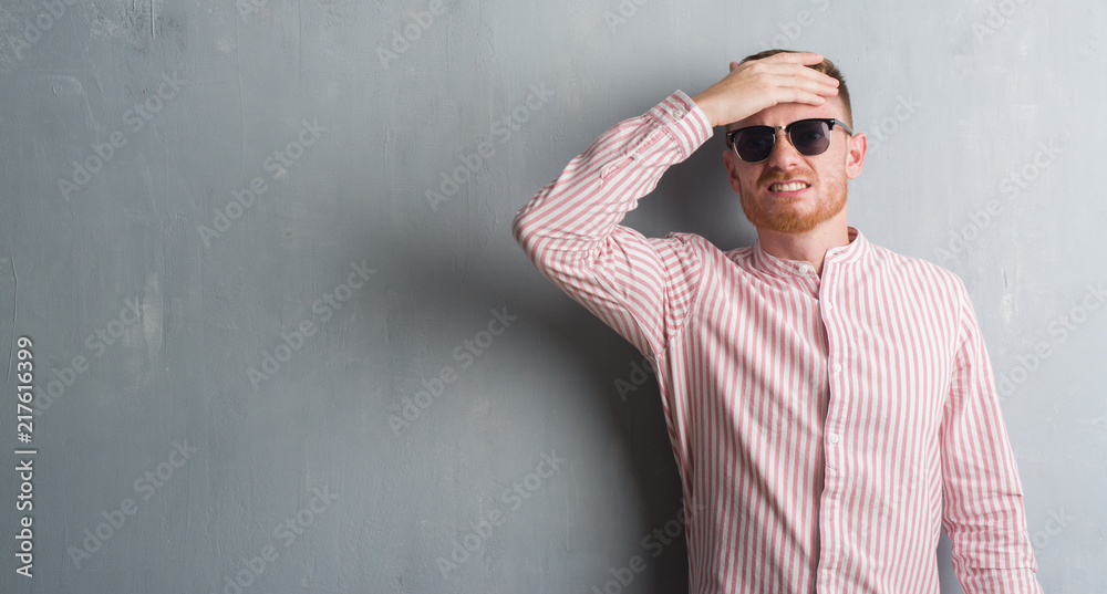 Young redhead man over grey grunge wall wearing sunglasses stressed with hand on head, shocked with shame and surprise face, angry and frustrated. Fear and upset for mistake. Young redhead man over grey grunge wall wearing sunglasses stressed with hand on head, shocked with shame and surprise face, angry and frustrated. Fear and upset for mistake.
