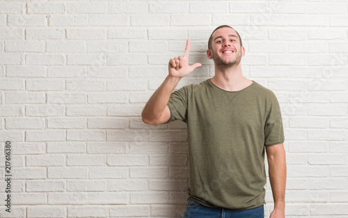 Young caucasian man standing over white brick wall showing and pointing up with fingers number two while smiling confident and happy.