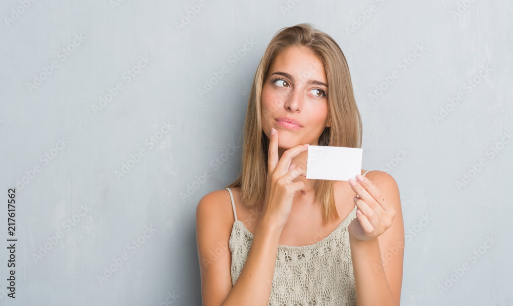 Beautiful young woman over grunge grey wall holding blank visit card serious face thinking about question, very confused idea Beautiful young woman over grunge grey wall holding blank visit card serious face thinking about question, very confused idea