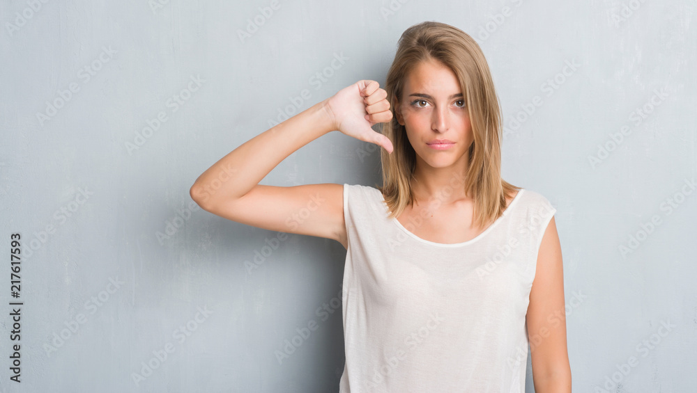 Beautiful young woman standing over grunge grey wall with angry face, negative sign showing dislike with thumbs down, rejection concept Beautiful young woman standing over grunge grey wall with angry face, negative sign showing dislike with thumbs down, rejection concept
