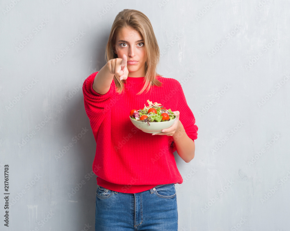 Beautiful young woman over grunge grey wall eating tomato salad pointing with finger to the camera and to you, hand sign, positive and confident gesture from the front
