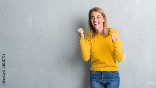 Beautiful young woman standing over grunge grey wall very happy and excited doing winner gesture with arms raised, smiling and screaming for success. Celebration concept.