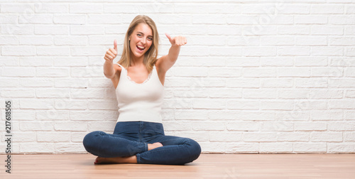 Beautiful young woman sitting on the floor at home approving doing positive gesture with hand, thumbs up smiling and happy for success. Looking at the camera, winner gesture.