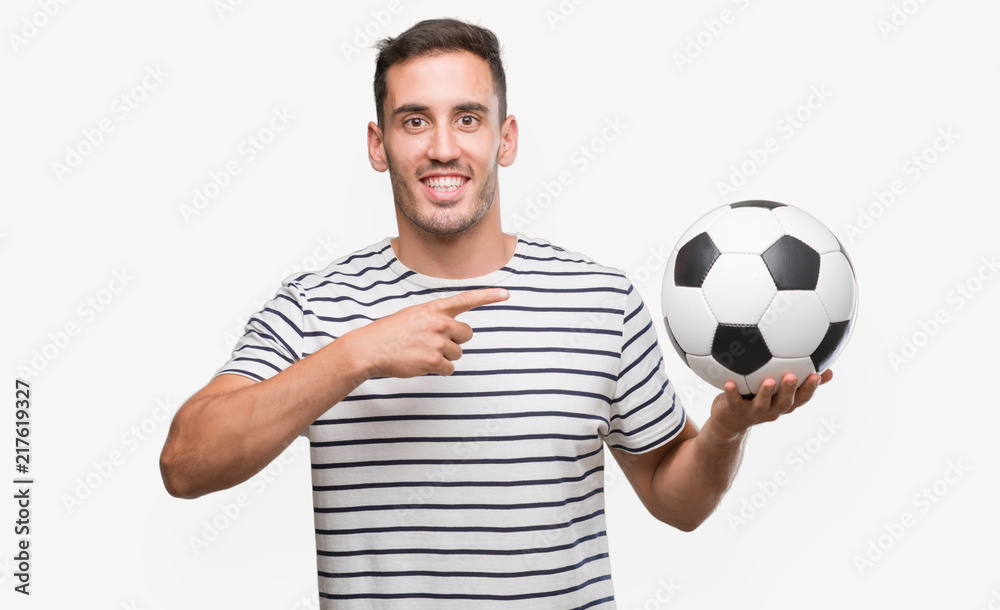 Handsome young man holding soccer football very happy pointing with hand and finger Handsome young man holding soccer football very happy pointing with hand and finger