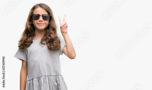 Brunette hispanic girl wearing sunglasses showing and pointing up with fingers number two while smiling confident and happy.