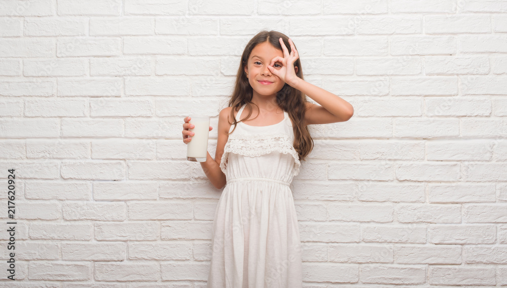 Young hispanic kid over white brick wall drinking a glass of milk with happy face smiling doing ok sign with hand on eye looking through fingers Young hispanic kid over white brick wall drinking a glass of milk with happy face smiling doing ok sign with hand on eye looking through fingers