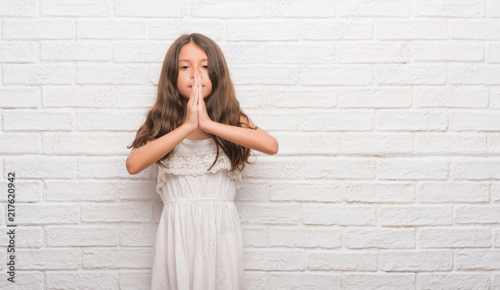 Young hispanic kid over white brick wall praying with hands together asking for forgiveness smiling confident.