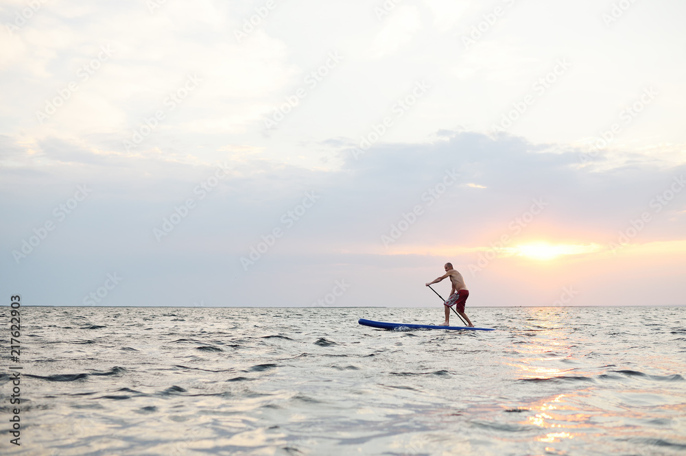 A man stands on a SUP board against the background of the sea and the sunset.