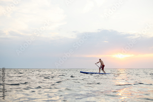 A man stands on a SUP board against the background of the sea and the sunset. © Evgeniy Kalinovskiy
