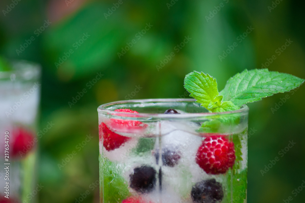 Cold lemonade with lemon, mint and berries in a transparent glass with facets. On an old wooden table. In the background, the green foliage is a soft blurred focus. Cold lemonade with lemon, mint and berries in a transparent glass with facets. On an old wooden table. In the background, the green foliage is a soft blurred focus.