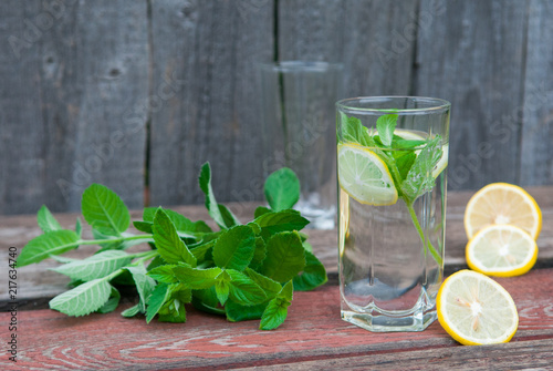 Water with lemon and mint in a transparent glass with facets. On an old wooden table. In the background, the green foliage is a soft blurred focus.